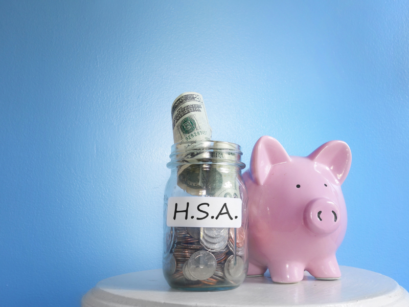 A glass jar labeled HSA containing coins and a rolled dollar bill next to a pink piggy bank, symbolizing health savings account budgeting and financial planning.